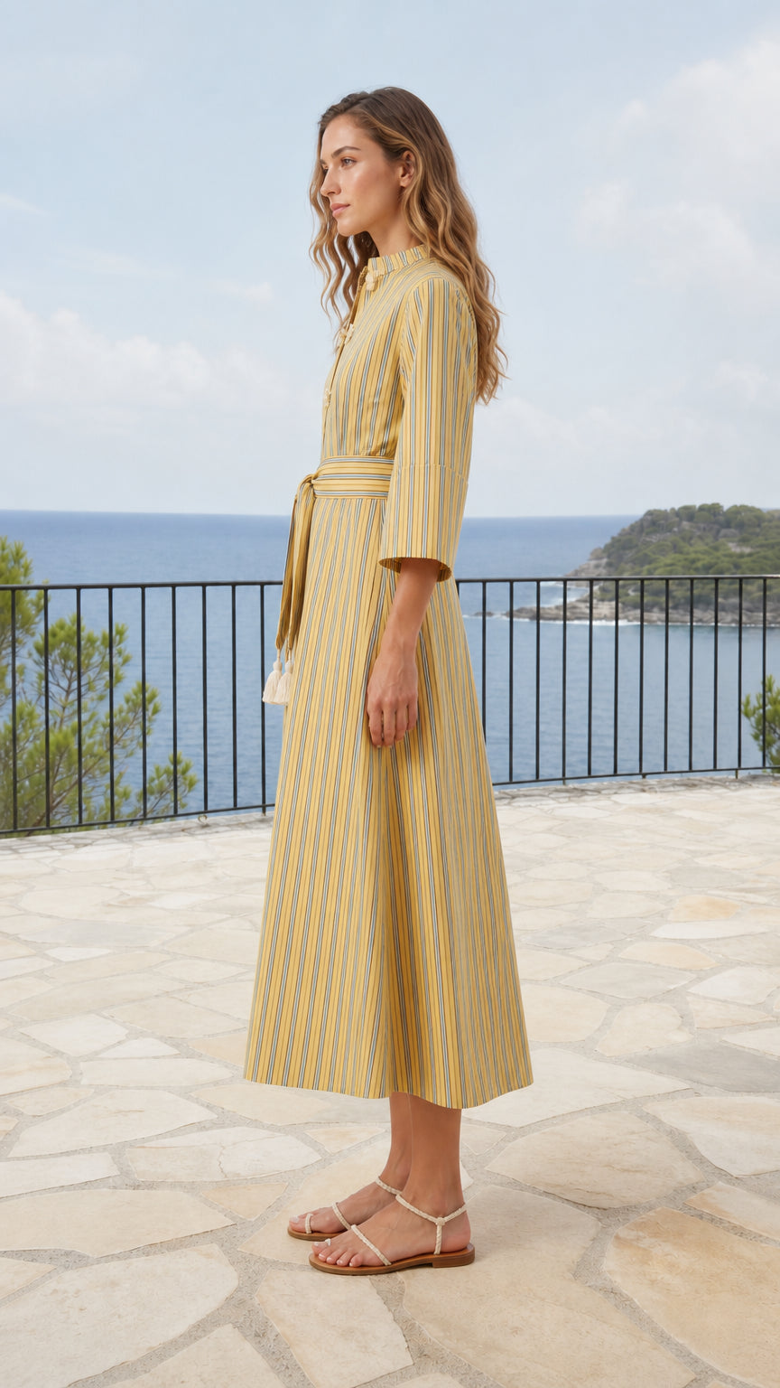 Side profile of model wearing yellow striped A-line midi dress on seaside balcony