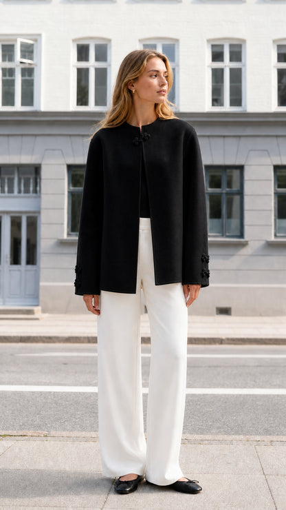 Front full view of woman wearing black collarless knot clasp jacket with white trousers